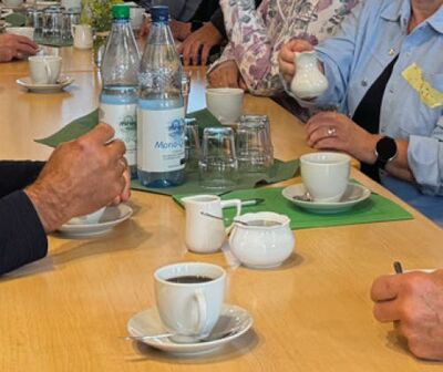 Man sieht eine gedeckte Kaffeetaffel, an der mehrere Personen sitzen und gerade Kaffee trinken.