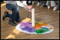Eine hohe Kerze steht auf einer geschmückten Unterlage. Jemand zündet dir Kerze gerade an. Die anderen Teilnehmenden der Andacht sitzen in einem großen Stuhlkreis drumherum.