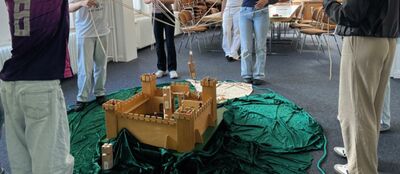 Man sieht eine Burg aus Holz auf dem Boden. Eine Gruppe Konfirmanden stehen um die Burg herum und müssen mit Seilen gemeinsam einen Gegenstand in der Burg ablegen.